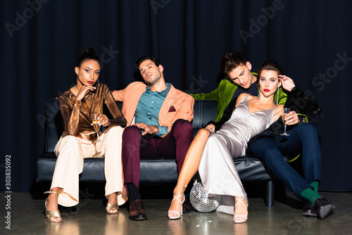 beautiful multicultural women and handsome men holding champagne glasses while sitting on sofa on dark blue