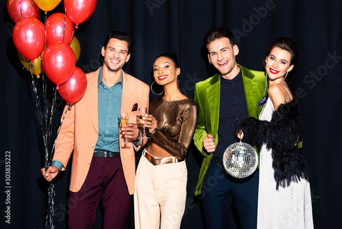 happy multicultural women and handsome men holding champagne glasses near balloons on dark blue
