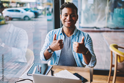 Half length portrait of handsome funny emotional hindu male person showing thumbs up while sitting at table in coffee shop.Cheerful mature man looking at camera during making fun gesturing indoors