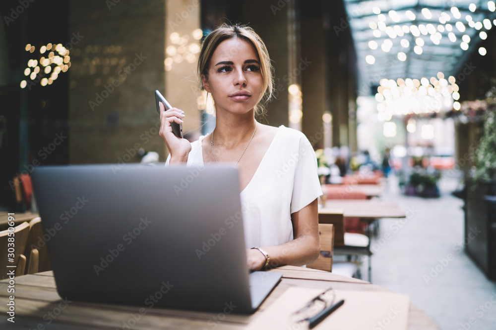 Beautiful caucasian hipster girl holding smartphone gadget in hand and waiting for call from colleague while sitting with laptop in cafeteria, young skilled woman spending time with netbook indoors