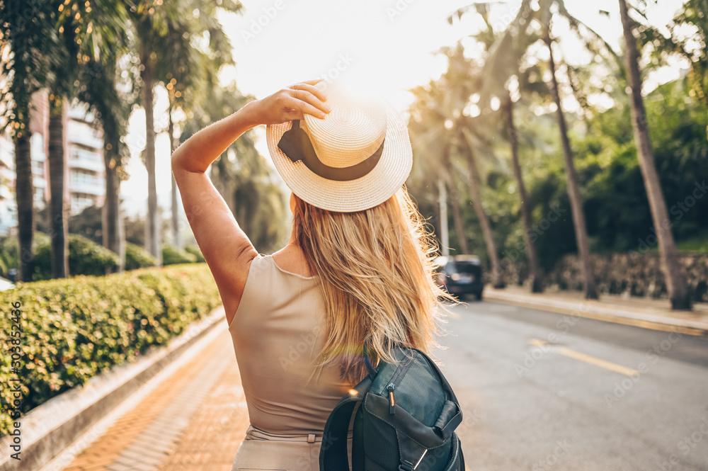 Chinese tourist walking in city streets on Asia vacation tourism. Caucasian beautiful woman in hat with backpack travel lifestyle concept. Walking street, green palms and highway in Sanya, Hainan.  - obrazy, fototapety, plakaty 