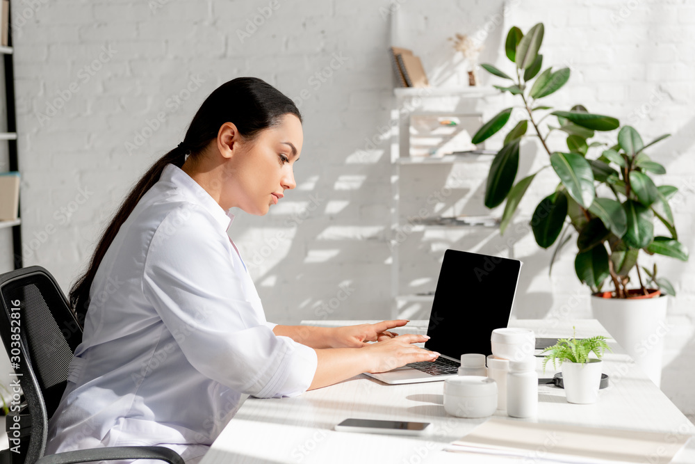 attractive dermatologist sitting at table and using laptop in clinic