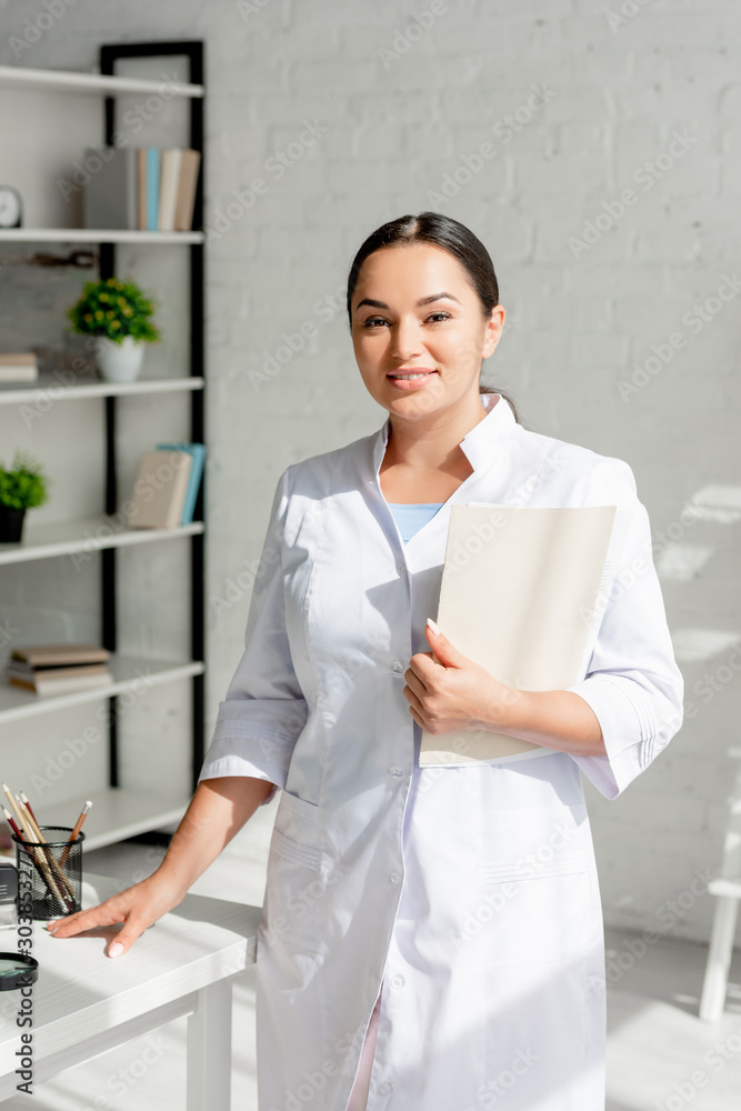 smiling dermatologist holding folder and looking at camera in clinic smiling dermatologist holding folder and looking at camera in clinic