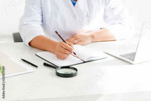 cropped view of dermatologist sitting at table and writing in notebook in clinic