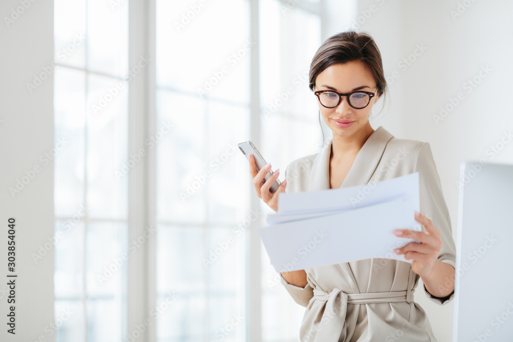 Satisfied female employee focused in documents, reads prepared report attentively, holds modern smartphone, wears transparent glasses and business suit poses over office interior checks monthly income Satisfied female employee focused in documents, reads prepared report attentively, holds modern smartphone, wears transparent glasses and business suit poses over office interior checks monthly income