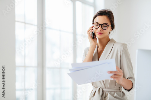 Horizontal shot of business lady discusses details of contract, works in office, concentrated on information from accountings, holds paper documents during telephone conversation, dressed formally
