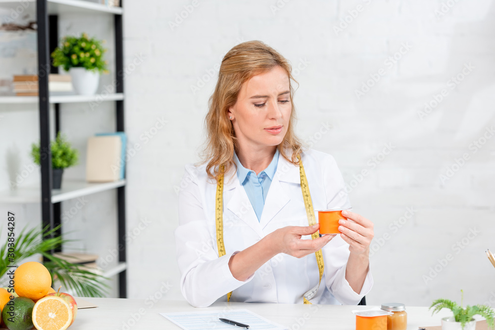 attractive nutritionist sitting at table and holding yogurt in clinic attractive nutritionist sitting at table and holding yogurt in clinic