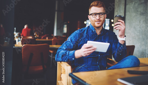 Pondering hipster guy holding digital tablet in hand and thinking about next publication at own web blog, caucasian man using 4g wireless connection on modern touch pad for communication online