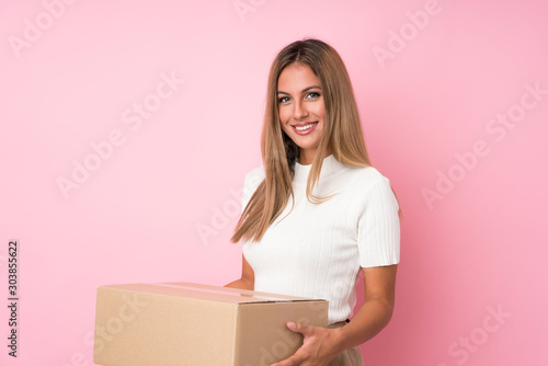 Young blonde woman over isolated pink background holding a box to move it to another site © luismolinero