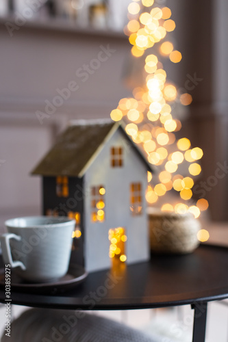 Closeup blurred photo of lighted from inside christmas decorative house and two cups standing on black table