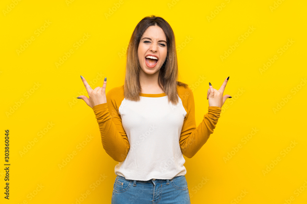 Pretty young woman over isolated yellow wall making rock gesture Pretty young woman over isolated yellow wall making rock gesture