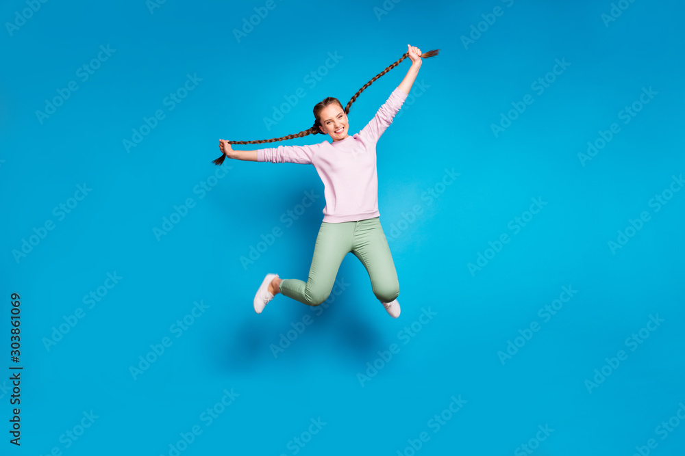 Full length photo of positive cheerful millennial girl jump hold her braids have fun wear casual style outfit white sneakers isolated over blue color background Full length photo of positive cheerful millennial girl jump hold her braids have fun wear casual style outfit white sneakers isolated over blue color background