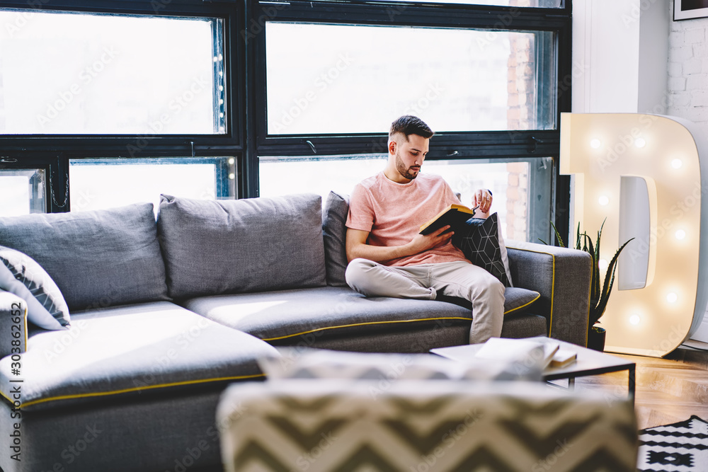 Pensive young man resting at home on comfortable sofa and reading book in modern apartment with stylish interior.Hipster guy enjoying literature plot sitting on cozy couch in design flat