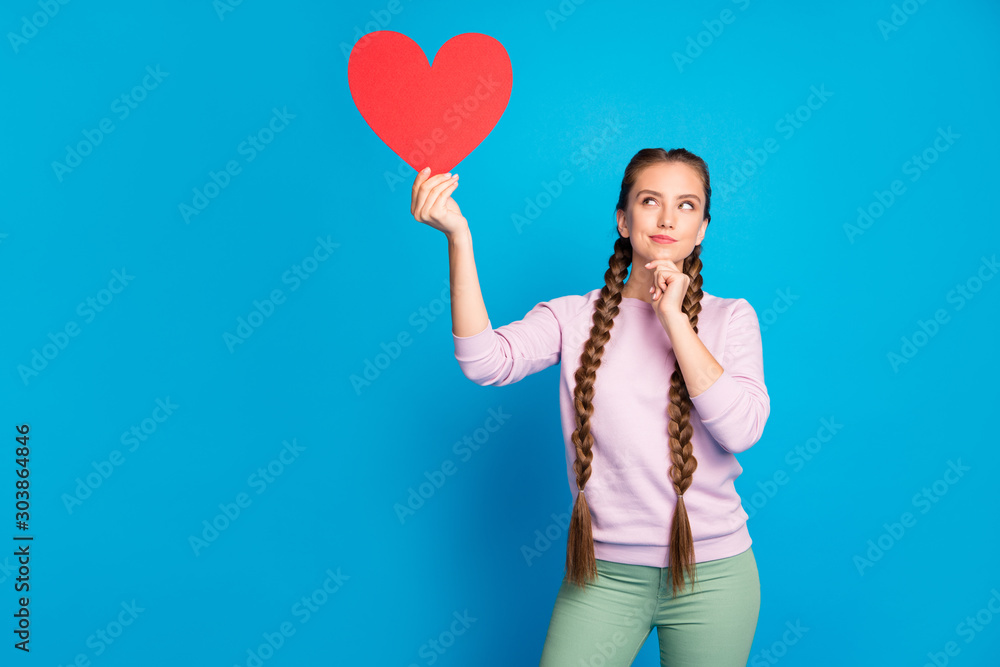 Portrait of thoughtful interested youth girl with long pigtails hold red paper card heart gift for valentine day think about cupid date wear pink pullover green pants isolated blue color background Portrait of thoughtful interested youth girl with long pigtails hold red paper card heart gift for valentine day think about cupid date wear pink pullover green pants isolated blue color background