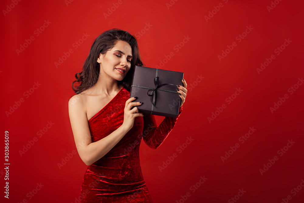 Obraz premium girl woman on a red background in a red fashion dress holds a black gift in hands. Happy Birthday. merry christmas, new year,&nbsp; valentines day. happiness youth beauty femininity. place for text copy