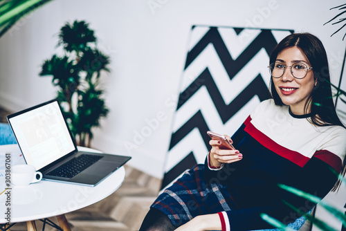 Happy female blogger in optical spectacles holding smartphone in hand and looking away with cute smile on face while sitting in stylish home interior near desktop with laptop device with mock up area