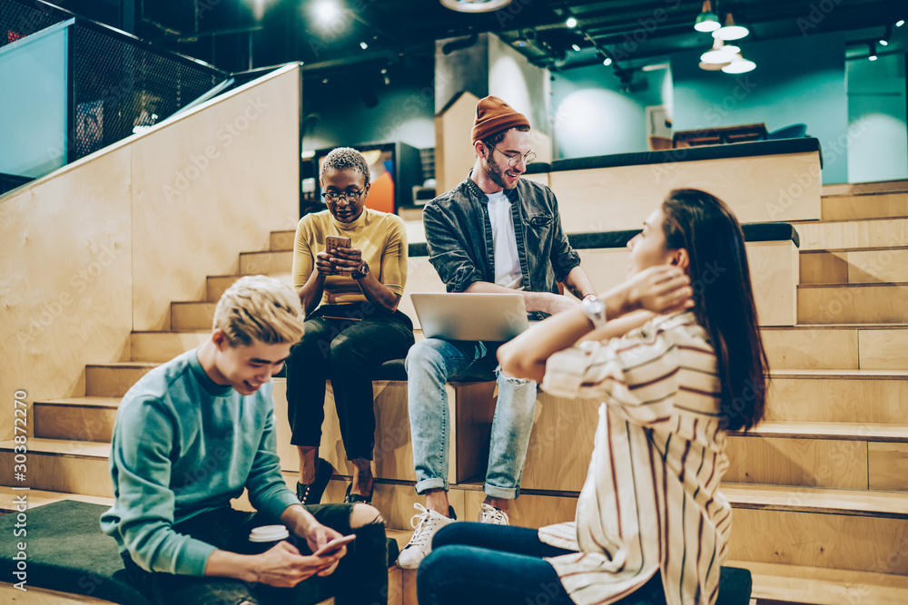 Fototapeta premium Positive multiracial colleagues communicating during free time on break in campus using modern gadgets, group of male of female hipsters spending time together share creative ideas and networking.