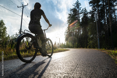 Woman riding a bike on a bicycle path next to a pine forest, under the beginning of warm, summer rain and sun.