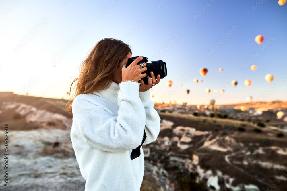 A tourist photographer girl wearing white sweater on a mountain top enjoying wonderful view and taking a picture of balloons in Cappadocia A tourist photographer girl wearing white sweater on a mountain top enjoying wonderful view and taking a picture of balloons in Cappadocia