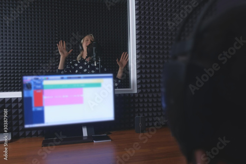 Woman professional vocalist singer in headphones singing in microphone and sound engineer in studio recording song. Creating new melody audio composition on musical session at recording company.