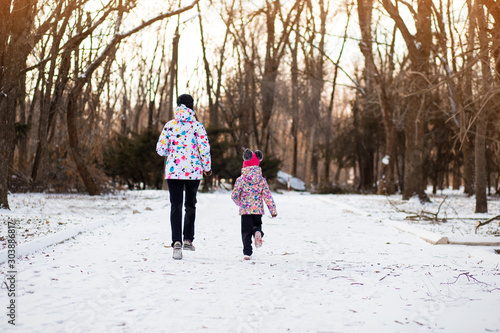 Mother and little girl in colored jackets jogging by snow in winter park. Concept of instill sports health habits in children