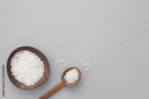 White bath salt in bowl and spoon from coconut shell on gray background. Natural spa products concept. Top view, flay lay, copy space