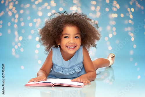 childhood, school and education concept - happy smiling little african american girl reading book over festive lights on blue background