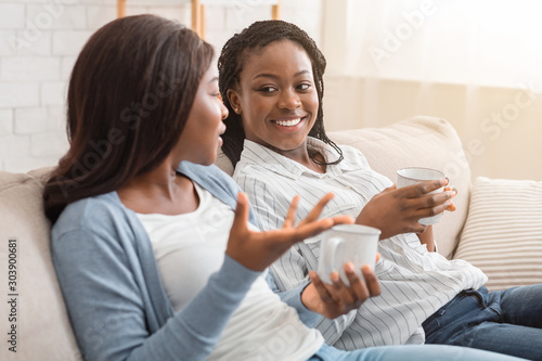 Two black girls sitting together on sofa, chatting and drinking tea