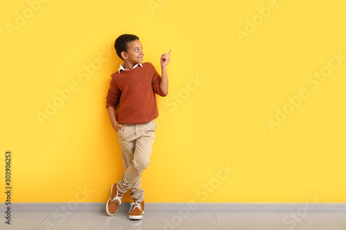 Fashionable African-American boy in autumn clothes pointing at something while standing near color wall