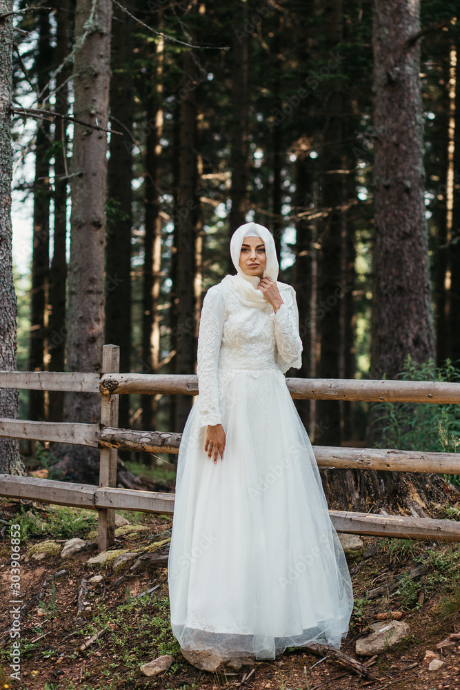 Beautiful elegant bride with hijab in lace wedding dress with long full skirt and long sleeves. Outdoors, in the woods, wooden fence in the background.