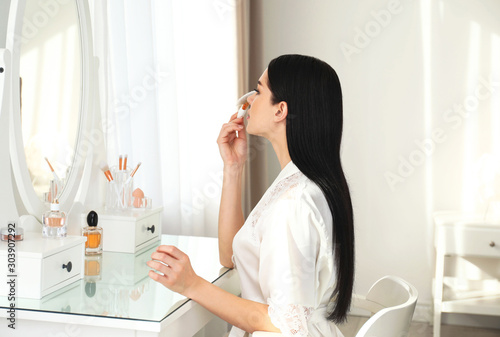 Beautiful young woman in robe doing make up at dressing table indoors