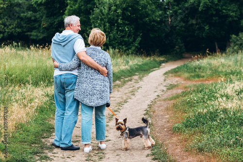 mature couple is walking with dog in park. Elderly couple resting in nature with a dog. Full-length portrait of an elderly man and woman in white shirts and jeans. Stylish and modern grandparents.
