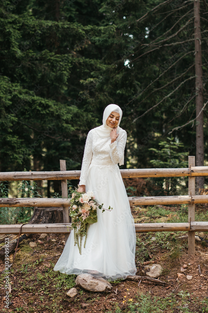 Fototapeta premium Beautiful elegant bride with hijab in lace wedding dress with long full skirt and long sleeves. She is holding a big bouquet of flowers. Outdoors, in the woods.
