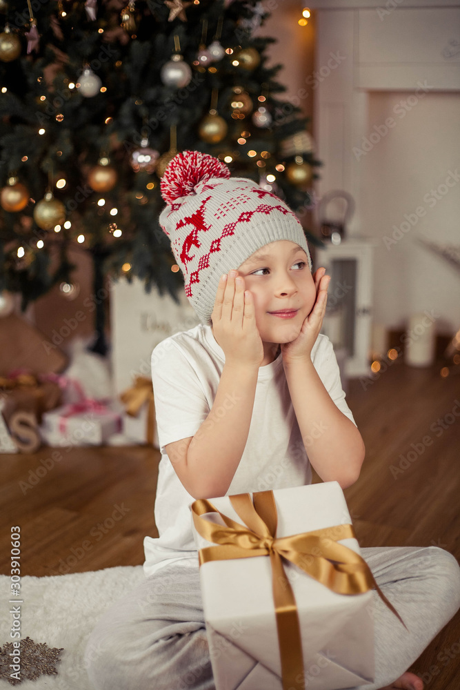 Little cute boy 5 years old in a Christmas hat opens a gift near the Christmas tree. Christmas. New Year. Little cute boy 5 years old in a Christmas hat opens a gift near the Christmas tree. Christmas. New Year.