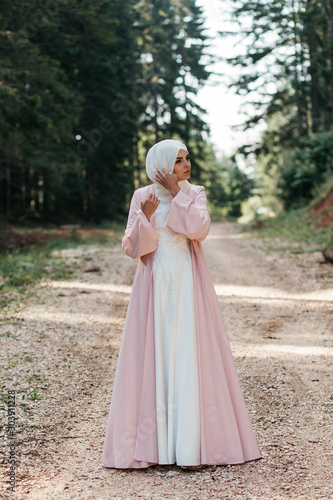 Beautiful elegant bride with hijab in nude pink dress with long full skirt and long sleeves. Outdoors, on te road. She is posing for fashion clothes. 