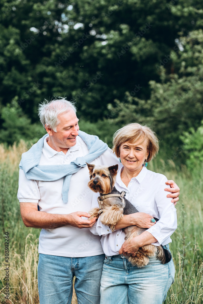 A mature couple is walking with a dog in a park. Elderly couple resting in nature with a dog. Close-up portrait of an elderly man and woman in white shirts and jeans. Stylish and modern grandparents. A mature couple is walking with a dog in a park. Elderly couple resting in nature with a dog. Close-up portrait of an elderly man and woman in white shirts and jeans. Stylish and modern grandparents.