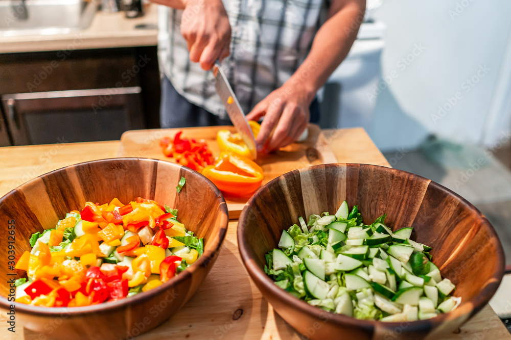 Man chopping vegetables on cutting board table with two large bowls of fresh vegan salad with red orange bell peppers in kitchen