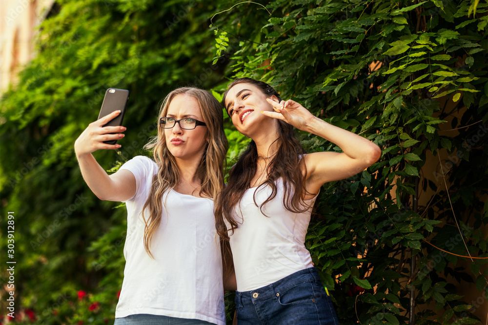Φωτογραφία Two funny young girls are taking selfie photos on the smartphone near the wall o