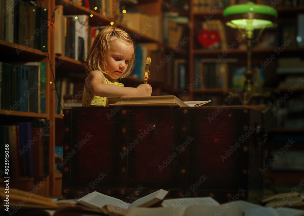 A little girl at home library reading a book sitting in an old chest holding a magnifying glass. Children and education many books on the floor, night time A little girl at home library reading a book sitting in an old chest holding a magnifying glass. Children and education many books on the floor, night time