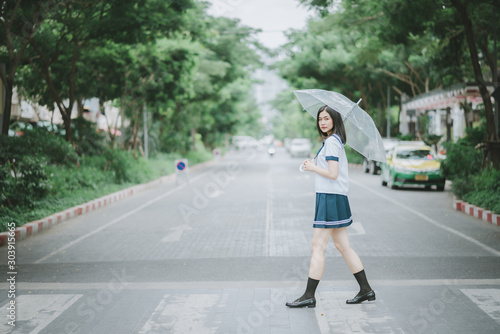 pretty asian girl in Japanese student uniform with short dark hair and holding an umbrella turning to look at camera while walking across the street with a lot of trees along the street