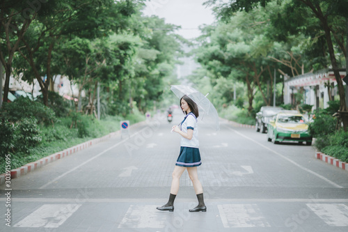 pretty asian girl in Japanese student uniform with short dark hair holding an umbrella and walking across the street in the middle of somewhere with a lot of trees along the street