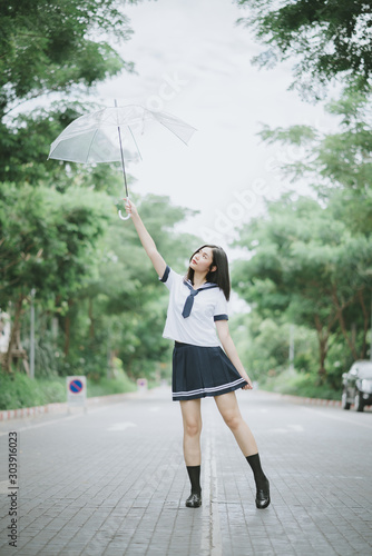 asian lady with short straight hair in Japanese student uniform holding a transparent umbrella with arm raised looking up to the umbrella posing on the street standing with pointed legs