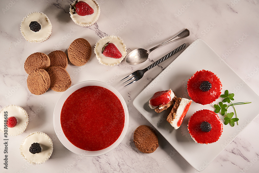 Φωτογραφία Top view of homemade cheesecakes dipped in strawberry sauce and accompanied with homemade strawberry sauce, cookies, raspberries, blackberries and clover leaves on marble background