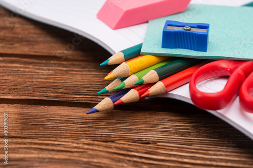 Colored pencils, sharpener, scissors and sheets for notes in an open notebook on a dark brown wooden surface. Flat lay. Back to school. Education. Office tool. Empty place for text.