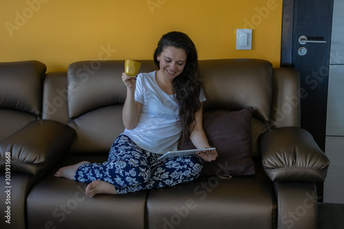 Beautiful latin woman sitting on the couch drinking coffee and checking her tablet