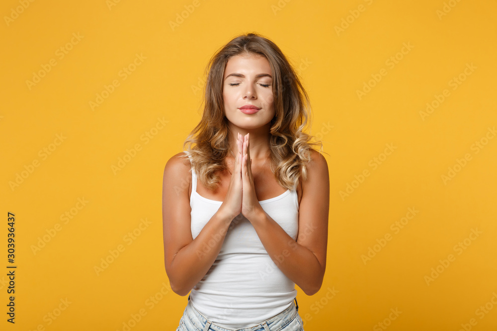 Young woman girl in light casual clothes posing isolated on yellow orange wall background in studio. People lifestyle concept. Mock up copy space. Holding hands folded in prayer, keeping eyes closed. Young woman girl in light casual clothes posing isolated on yellow orange wall background in studio. People lifestyle concept. Mock up copy space. Holding hands folded in prayer, keeping eyes closed.