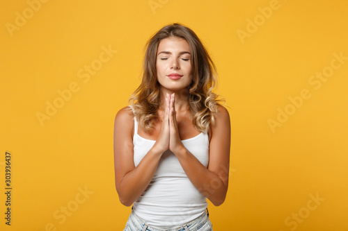 Young woman girl in light casual clothes posing isolated on yellow orange wall background in studio. People lifestyle concept. Mock up copy space. Holding hands folded in prayer, keeping eyes closed.