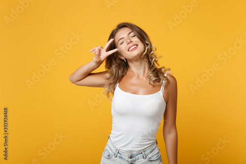 Funny young woman in light casual clothes posing isolated on yellow orange wall background, studio portrait. People lifestyle concept. Mock up copy space. Keeping eyes closed, showing victory sign.