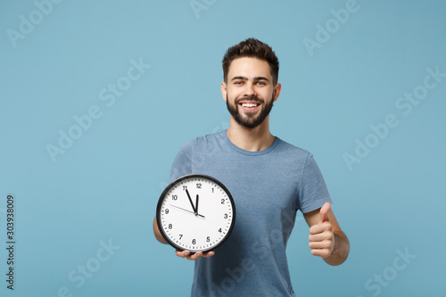 Young smiling handsome man in casual clothes posing isolated on blue background, studio portrait. People sincere emotions lifestyle concept. Mock up copy space. Hold in hands clock, showing thumb up.