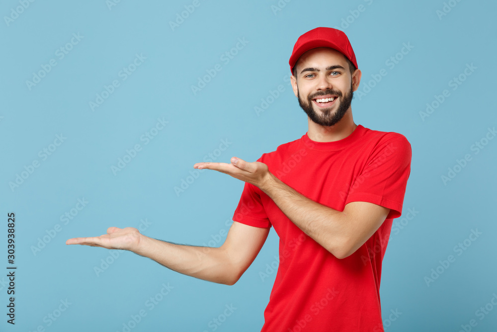Delivery man in red uniform workwear isolated on blue wall background, studio portrait. Professional male employee in cap t-shirt print working as courier dealer. Service concept. Mock up copy space. Delivery man in red uniform workwear isolated on blue wall background, studio portrait. Professional male employee in cap t-shirt print working as courier dealer. Service concept. Mock up copy space.